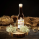 Eine Flasche mit dunkelrotem Holunderlikör davor ein Kristallglas gefüllt mit Likör umgeben von Holunderblüten auf einem Holzbrett auf dunklem Holzhintergrund